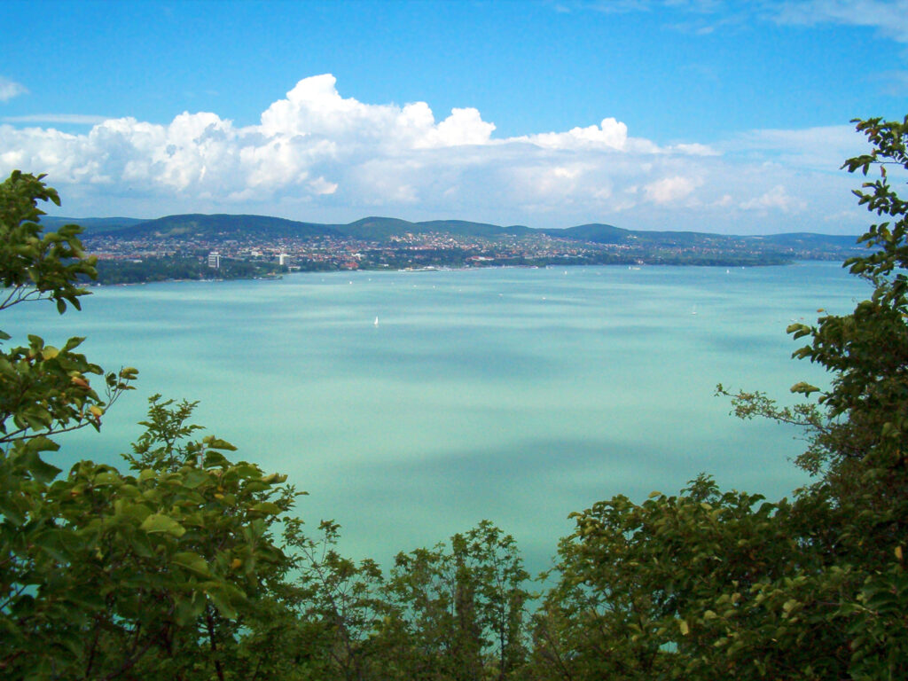 Vue panoramique sur le lac Balaton avec ses eaux turquoise entourées de collines verdoyantes et d’un ciel dégagé.